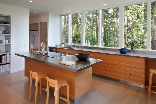 Просторна кухня с голям панорамен прозорец, покриващ почти цялата стена. Перваза на перваза на плота изглежда много органична в тази опция за дизайн на пространството.