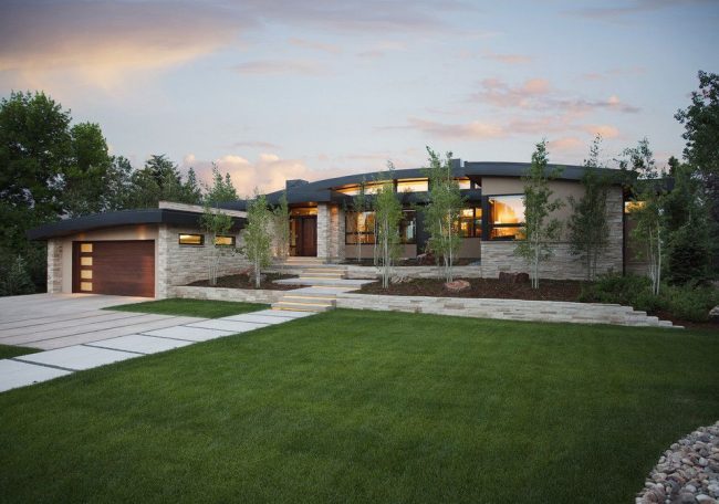 Модерен екстериор на къща с гараж: каменна облицовка, извит покрив, ярко осветление, зелена прилежаща зона