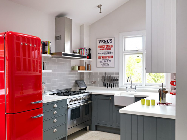 Šedá kuchyňská zástěra s imitací zdiva, šedá sada s bílou deskou, kovový lesk domácích spotřebičů - jednoduchý a funkční design, který zdůrazňuje jasné cákance. Červená retro chladnička je jedinečným prvkem v interiéru kuchyně