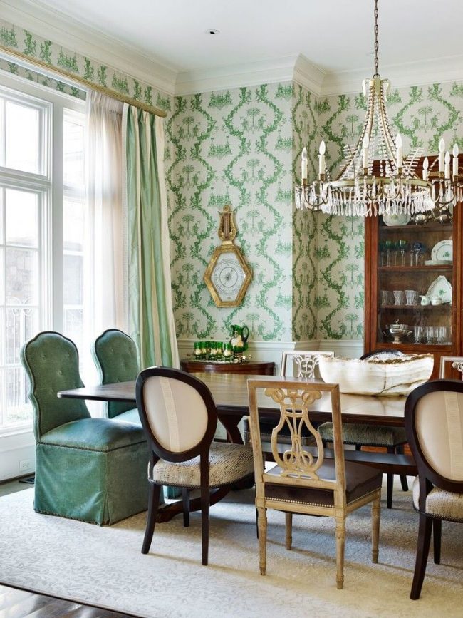 Klasická jídelna s dřevěným příborníkem na nádobí, křišťálovým lustrem, židlemi různých tvarů a textilními potahy, bílá tapeta se zelenými ornamenty